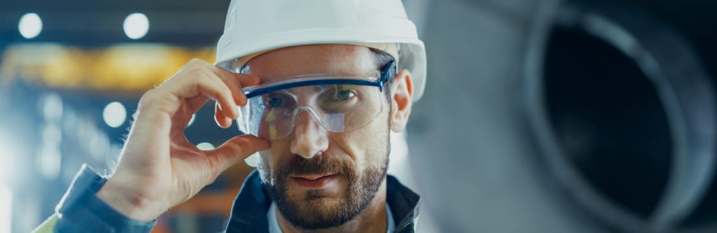 Photographie d'un chef d'équipe dans l'industrie, prise de face. Il tient ses lunettes de protection avec sa main et porte un casque de protection blanc.