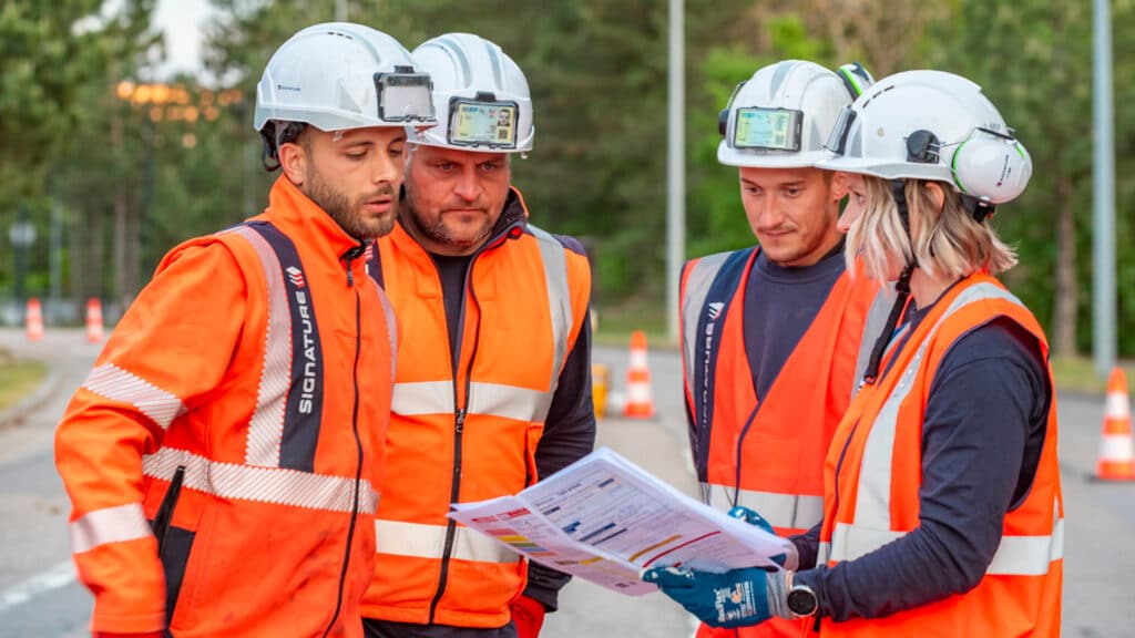 4 personnes se tiennent sur une route, en arrière plan, on peut voir forêt et des plots de chantiers oranges et blancs qui suivent la route. Les 4 personnes (3 hommes et 1 femme) portent leurs vêtements de sécurité (gilet orange fluo, casque, gants, casque anti-bruit), ils sont en train de regarder un manuel.
© Axel Heise