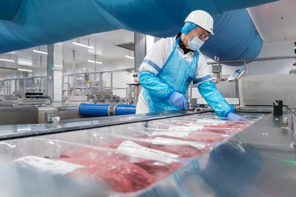 Un collaborateur d'Elivia travaille sur une ligne de conditionnement de viande. Sur la ligne, on peut voir les différents sachets défiler vers la prochaine étape. Le collaborateur porte une blouse, un tablier, des gants, des protèges avant-bras, une charlotte, un masque, des lunettes et un casque.