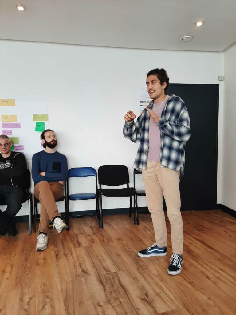 Le rappeur Paco se tient au milieu d'une pièce, il est en train d'expliquer les techniques à utiliser pour composer un rap. Au fond de la pièce, 4 chaises sont disposées. 2 d'entre elles sont occupées par des membres des équipes de voestalpine.